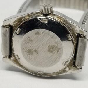 May include: Close-up of a vintage silver-toned wristwatch. The watch has a round, textured back with the text "MRG Stainless" and other markings. The watchband is a silver-toned, expandable metal band.