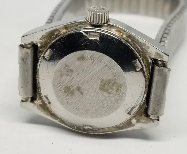 May include: Close-up of a vintage silver-toned wristwatch. The watch has a round, textured back with the text "MRG Stainless" and other markings. The watchband is a silver-toned, expandable metal band.