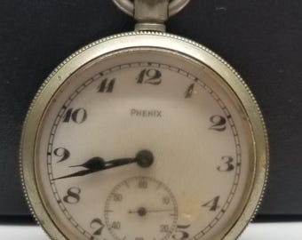 Puede incluir: Un reloj de bolsillo plateado con esfera blanca y manecillas negras. La esfera del reloj tiene números negros y la marca "PHENIX". Una esfera más pequeña está en la parte inferior. El reloj tiene un lazo plateado en la parte superior.