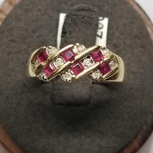 May include: A gold ring featuring a series of square-cut red gemstones and small, round, clear gemstones. The ring has a woven design, with the gemstones set in a diagonal pattern. The ring is displayed on a dark gray surface.