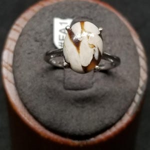 May include: A silver-toned ring featuring an oval-shaped stone with a smooth, polished surface. The stone displays a marbled pattern of creamy white and dark brown veins. The setting includes four small prongs securing the stone to the band.