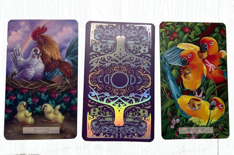 May include: Three tarot cards with colorful illustrations. The first card shows a rooster and hen with chicks, and the text "Ten of Cups". The second card has a purple background with a gold and silver tree design. The third card shows two parrots with cups, and the text "Three of Cups".
