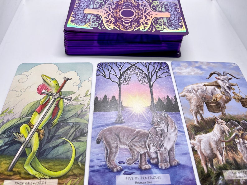 May include: Three tarot cards with colorful illustrations. The first card shows a green lizard holding a sword, with the text "Page of Swords". The second card shows two lynx in a snowy forest with the text "Five of Pentacles". The third card shows two white goats with a basket, with the text "Six of Pentacles".