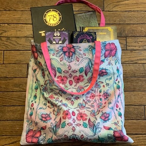 May include: A pink and white tote bag with a floral pattern featuring hummingbirds and crystals. The bag has pink straps and is filled with several books, including one with the text "78 Nautical".