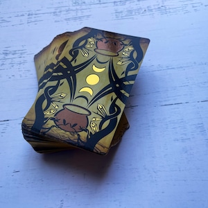 May include: A deck of tarot cards with a gold and black design. The top card shows a cauldron with a crescent moon and a full moon above it.