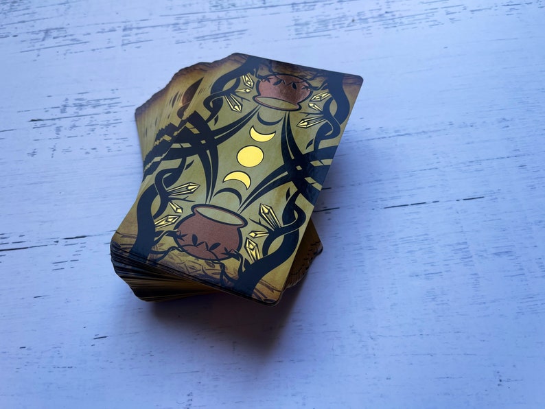 May include: A deck of tarot cards with a gold and black design. The top card shows a cauldron with a crescent moon and a full moon above it.