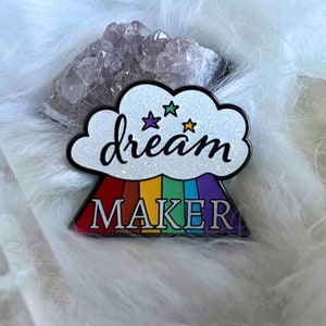 Puede incluir: Un pin de esmalte blanco con una nube de purpurina y el texto "dream maker" en escritura negra. La nube está sobre un arcoíris con un contorno negro. Hay tres pequeñas estrellas verdes en la nube.