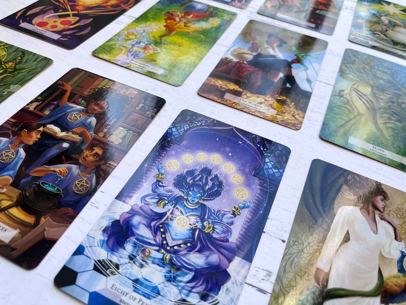 May include: A tarot card deck with colorful illustrations of people, animals, and symbols. The cards are laid out on a white surface. The card in the center shows a blue figure with a crown and a purple robe, surrounded by eight golden coins. The card text reads "Eight of Pentacles".
