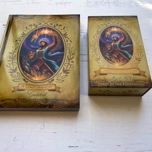 May include: A gold and brown tarot deck box with the text "Tarot Deck" and "Tarot of the Fantastical" on the front. The box is closed and has a gold trim. The box is sitting on a white wooden surface.