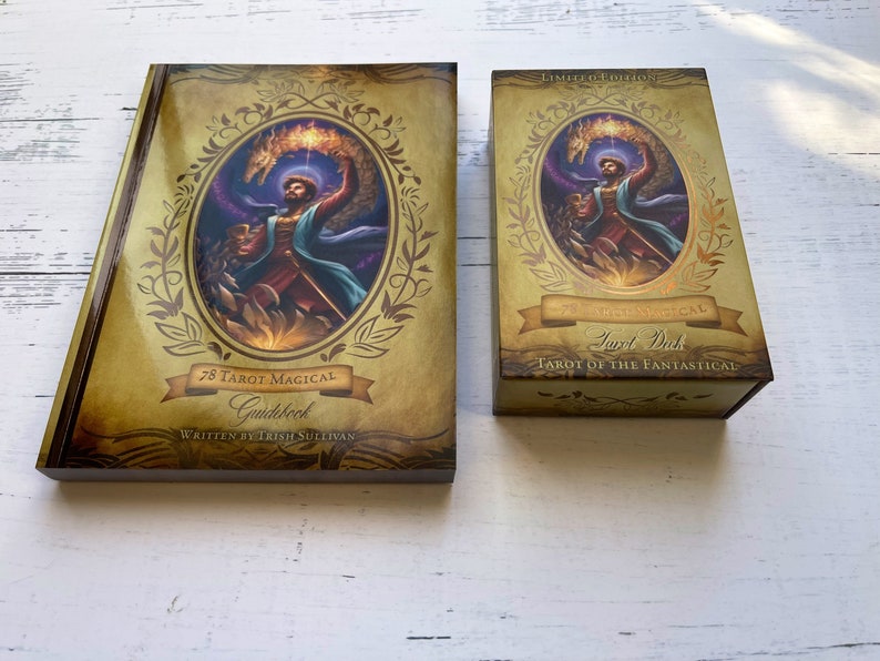 May include: A gold and brown tarot deck box with the text "Tarot Deck" and "Tarot of the Fantastical" on the front. The box is closed and has a gold trim. The box is sitting on a white wooden surface.