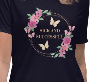 May include: A dark navy blue short-sleeve t-shirt featuring a central graphic of a circular gold frame adorned with pink flowers and light yellow butterflies. The text SICK AND SUCCESSFUL is printed in the center in a simple gold font.