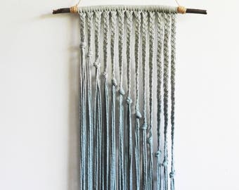 Op de afbeelding: Een wandhanger van lichtblauw katoenen koord opgehangen aan een donkere houten tak. Het ontwerp bestaat uit verticale gevlochten secties die overgaan in loshangende draden, waarbij elke vlecht op een andere lengte eindigt.