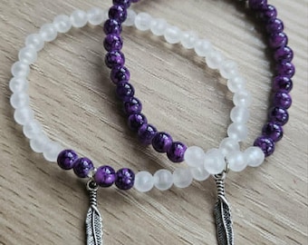 May include: Two stretch bracelets featuring round beads. One bracelet consists of deep purple marbled beads, while the other is made of translucent white frosted beads. Each bracelet includes a small, silver-toned metal feather charm pendant.