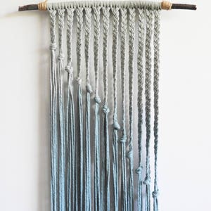 May include: A wall hanging made of light blue cotton cord suspended from a dark wooden branch. The piece features a series of vertical braided sections that transition into loose, hanging strands, each secured with a single knot at varying heights.