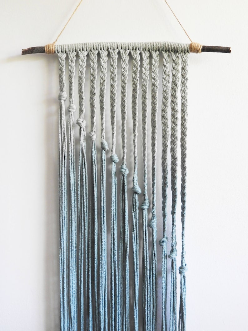 May include: A wall hanging made of light blue cotton cord suspended from a dark wooden branch. The piece features a series of vertical braided sections that transition into loose, hanging strands, each secured with a single knot at varying heights.