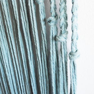 May include: A series of light blue cotton cords hanging vertically. The cords are arranged in a mix of loose, untwisted strands and sections featuring tight, braided patterns secured with simple overhand knots at various intervals.