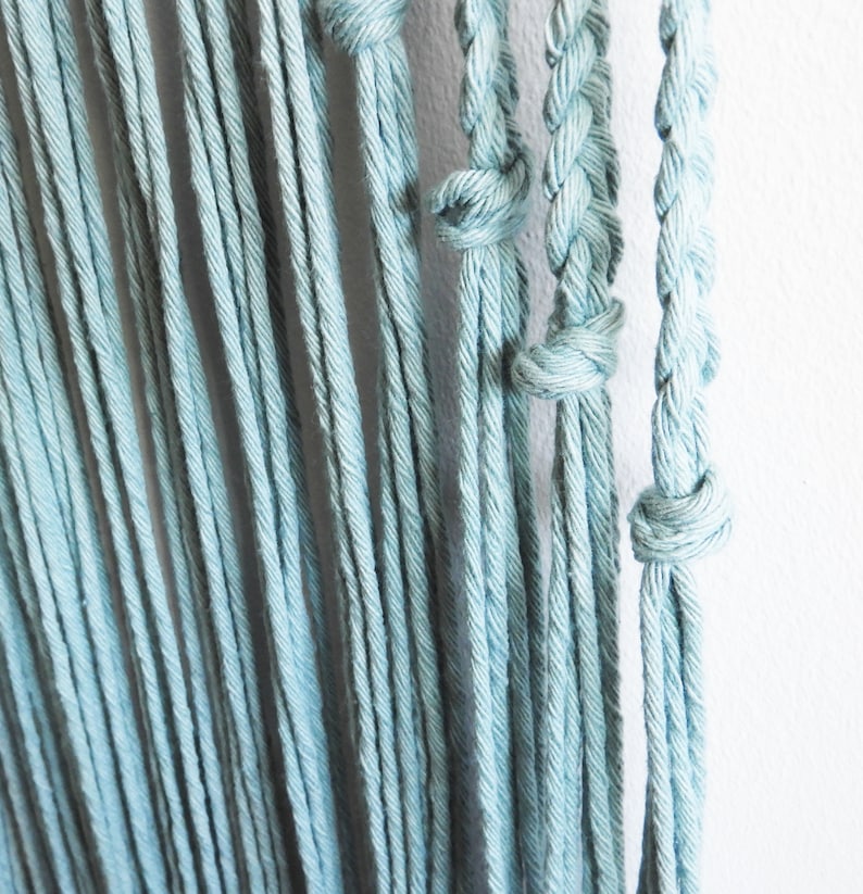 May include: A series of light blue cotton cords hanging vertically. The cords are arranged in a mix of loose, untwisted strands and sections featuring tight, braided patterns secured with simple overhand knots at various intervals.
