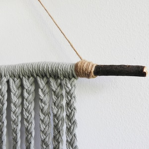 May include: A wall hanging featuring light gray cotton cord braided into vertical strands, suspended from a dark brown wooden branch. The cord is wrapped tightly around the branch with tan twine, and a thin tan string is attached for hanging.