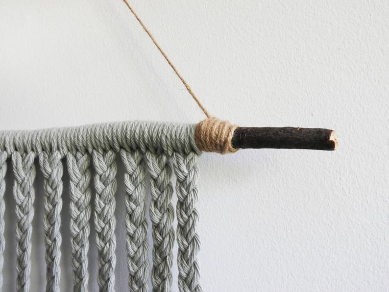 May include: A wall hanging featuring light gray cotton cord braided into vertical strands, suspended from a dark brown wooden branch. The cord is wrapped tightly around the branch with tan twine, and a thin tan string is attached for hanging.
