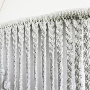 May include: A wall hanging featuring light gray cotton cord braided into multiple vertical strands. The cords are looped over a dark wooden rod, which is wrapped with natural tan jute twine at both ends and suspended by a thin string.