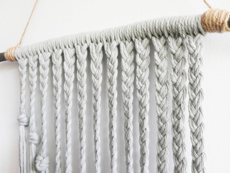 May include: A wall hanging featuring light gray cotton cord braided into multiple vertical strands. The cords are looped over a dark wooden rod, which is wrapped with natural tan jute twine at both ends and suspended by a thin string.