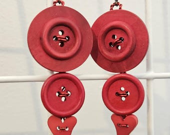 May include: A pair of dangling earrings featuring a vertical arrangement of two red circular buttons and one red heart-shaped button at the base. The components are connected by small metal rings and suspended from dark metal earring hooks.