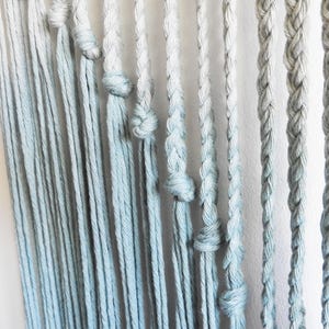 May include: A wall hanging featuring multiple vertical strands of cotton rope in a light blue and white ombre gradient. The strands vary in texture, with some left as loose, straight cords and others intricately braided or tied with simple knots.