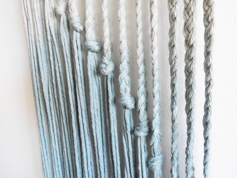 May include: A wall hanging featuring multiple vertical strands of cotton rope in a light blue and white ombre gradient. The strands vary in texture, with some left as loose, straight cords and others intricately braided or tied with simple knots.