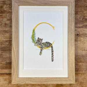May include: A framed art print with a watercolour illustration. A golden letter "G" is decorated with green fern and a resting clouded leopard. The artwork is set against a white background, framed in light wood.