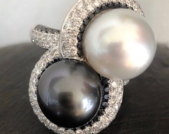 Pode incluir: Um anel de prata com duas pérolas, uma branca e uma preta, engastadas em um turbilhão de diamantes.