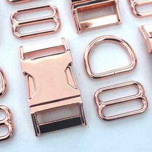 May include: Rose gold metal hardware assortment, including D-rings, buckles, and tri-glide slides. The shiny, metallic pieces are arranged on a white surface, ideal for crafting and accessories.