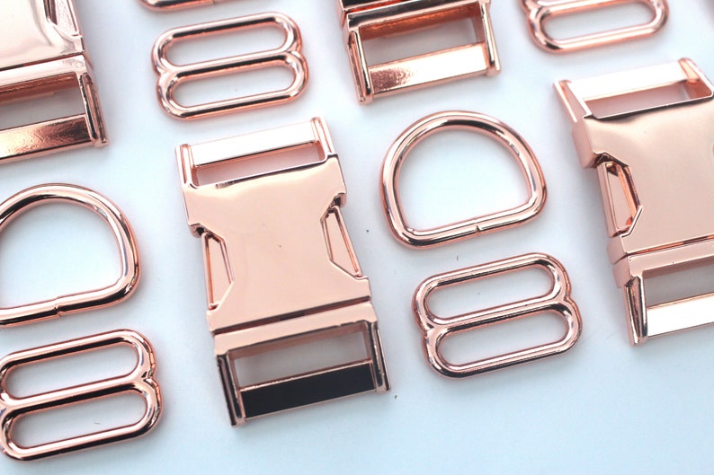 May include: Rose gold metal hardware assortment, including D-rings, buckles, and tri-glide slides. The shiny, metallic pieces are arranged on a white surface, ideal for crafting and accessories.