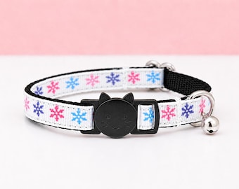 May include: A white pet collar featuring a repeating pattern of pink, blue, and purple snowflakes. The collar includes a black plastic buckle shaped like a cat face, a black nylon backing, a silver metal D-ring, and a small silver jingle bell.