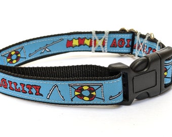 May include: A light blue pet collar with a black nylon backing and a black plastic side-release buckle. The fabric features a repeating pattern of agility course obstacles, including tires and seesaws, alongside the red text AGILITY.