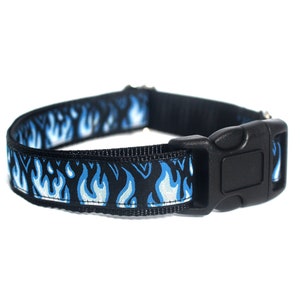 May include: Black dog collar with a blue flame pattern. The collar has a black plastic buckle and a metal loop for attaching a leash. The flame design is in shades of blue and white.