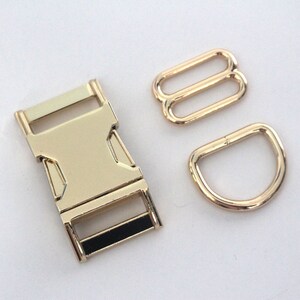May include: A collection of gold-coloured metal hardware components, including a buckle, two rectangular sliders, and a D-ring. The buckle features a black accent. These components are likely for crafting or bag making.
