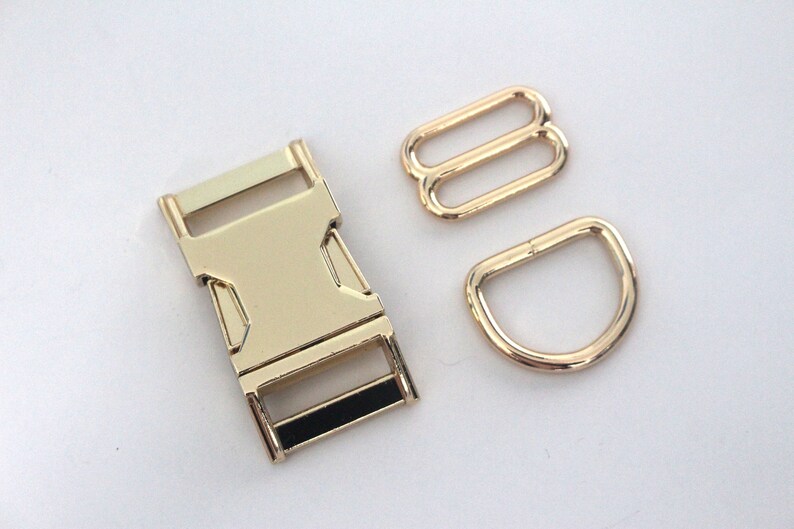 May include: A collection of gold-coloured metal hardware components, including a buckle, two rectangular sliders, and a D-ring. The buckle features a black accent. These components are likely for crafting or bag making.