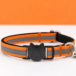 May include: An orange and grey reflective cat collar featuring a black plastic buckle shaped like a cat head with etched facial features. The collar includes a silver metal D-ring and a small silver bell attached to the side of the strap.