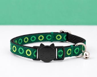May include: A dark green pet collar featuring a repeating pattern of light green four-leaf clovers. The collar includes a black plastic buckle shaped like a cat face, a silver metal D-ring, and a small silver bell attached to the side.