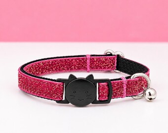 May include: A pet collar featuring a vibrant pink glittery fabric strap with a black nylon backing. The collar includes a black plastic breakaway buckle shaped like a cat face with etched whiskers and ears, along with a small silver metal bell.