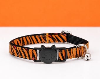 May include: A pet collar featuring an orange and black tiger stripe pattern. The collar includes a black plastic buckle shaped like a cat face with engraved whiskers and ears, a silver metal D-ring, and a small silver bell attached to the side.