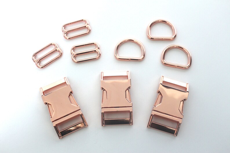 May include: Rose gold metal hardware set including D-rings, slide adjusters, and side-release buckles. The set is ideal for crafting dog collars, bag straps, or other DIY projects. The hardware has a shiny, metallic finish.