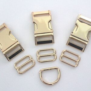 May include: A selection of gold-coloured metal hardware components, including three buckle clasps, two sets of double-loop slides, and a D-ring. The hardware is arranged on a white surface, suitable for various crafting projects.