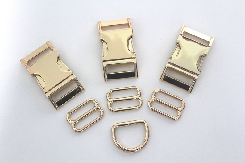 May include: A selection of gold-coloured metal hardware components, including three buckle clasps, two sets of double-loop slides, and a D-ring. The hardware is arranged on a white surface, suitable for various crafting projects.