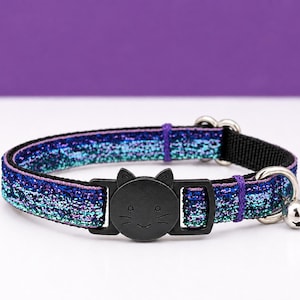 May include: A pet collar featuring a glittery blue, purple, and teal gradient fabric strap. The collar includes a black plastic breakaway buckle shaped like a cat face with etched whiskers and ears, along with a small silver metal jingle bell.