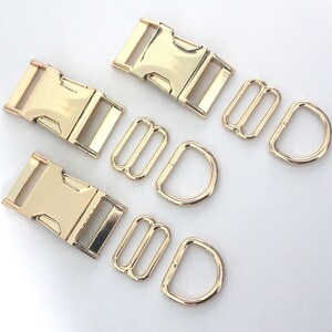 May include: A set of gold-coloured metal hardware, comprising two buckle clasps, two tri-glide slides, and two D-rings. The hardware is likely for crafting or bag making. The pieces are arranged on a white surface.