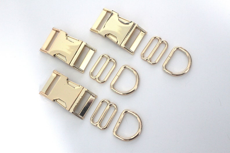 May include: A set of gold-coloured metal hardware, comprising two buckle clasps, two tri-glide slides, and two D-rings. The hardware is likely for crafting or bag making. The pieces are arranged on a white surface.