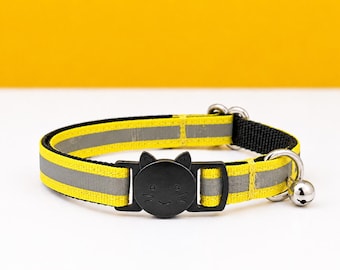 Könnte beinhalten: Ein leuchtend gelbes und schwarzes Katzenhalsband mit einem reflektierenden grauen Streifen in der Mitte. Das Halsband verfügt über eine schwarze Kunststoffschnalle in Katzenkopfform und eine kleine silberne Metallglocke an einem Ring an der Seite.