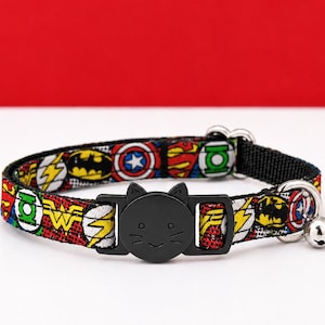 May include: A pet collar featuring a colourful comic book superhero pattern with various logos, including Batman, Wonder Woman, and Captain America. The collar includes a black plastic buckle shaped like a cat face and a small silver metal bell.