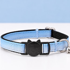 May include: A light blue and black cat collar with a black cat-shaped buckle. The collar has a silver bell and a metal ring. The collar is approximately 12 inches long.